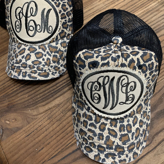 Two snapback hats with a camouflage pattern and a circular monogram design in the center, placed on a wooden surface.